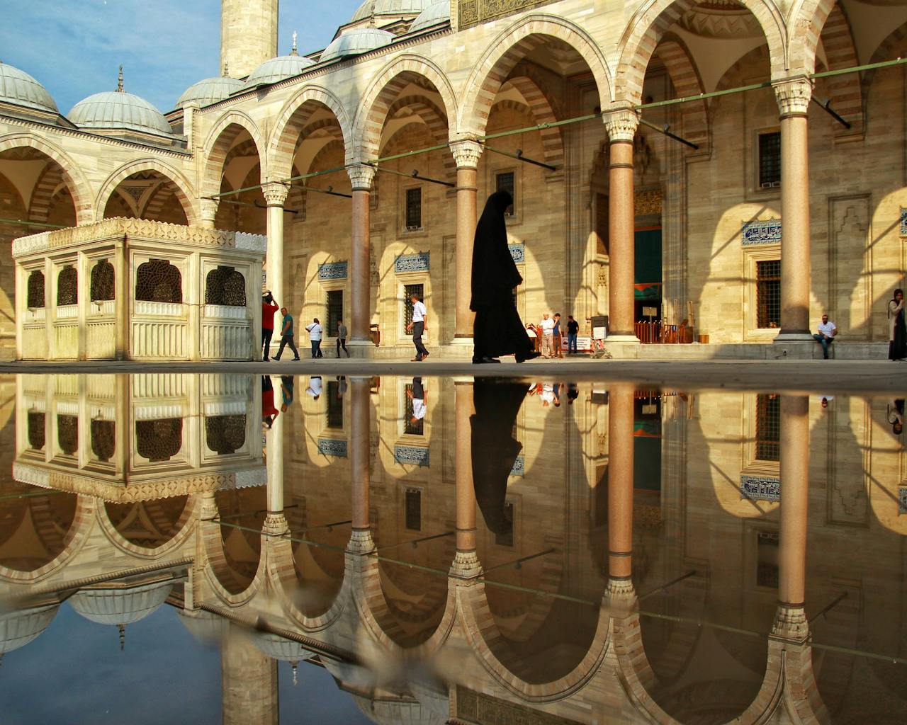 Silhouetted figure walks through Ottoman courtyard with arch reflections in water.